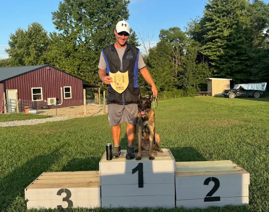 Joe Underwood standing on the first-place podium with his Belgian Malinois, holding a Ring 2 1st Place plaque